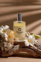 A bottle of Pentaura No. 75 perfume with a silver cap and cream-colored label, styled on a piece of driftwood with white plumeria flowers against a warm brown background with leaf shadows.