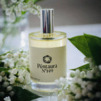 A clear glass perfume bottle with a reflective silver cap and a cream-colored label reading 'Péntaura N°149', styled with white flowers and green leaves around it