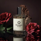 A bottle of Péntaura N°175 perfume centered in a styled studio setup, surrounded by red roses and a piece of driftwood against a dark brown background.
