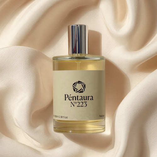 A clear glass perfume bottle with pale golden liquid, a cream label, and a reflective silver cap, centered on draped cream satin fabric. The label reads Péntaura No 223.