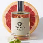 A bottle of Péntaura N°322 perfume centered in front of a sliced grapefruit, with a small white flower in the foreground on a clean white background.