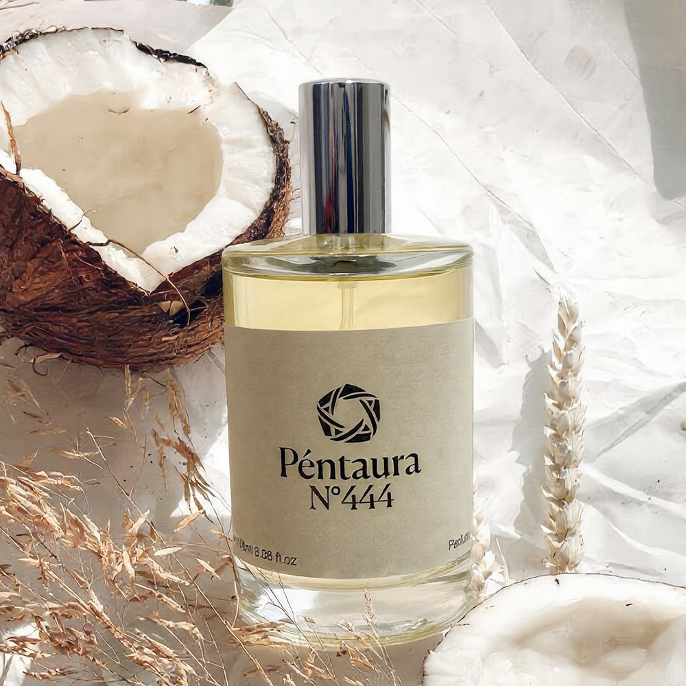 A bottle of Péntaura N°444 perfume with a silver cap and beige label, styled with a halved coconut, dried grass, and wheat on a white textured background.
