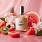 A bottle of Péntaura N°571 perfume centered on a glossy pink surface, surrounded by watermelon slices, kiwi, white flowers, and soft pink fabric. The bottle has a beige label with black text and a reflective silver cap.